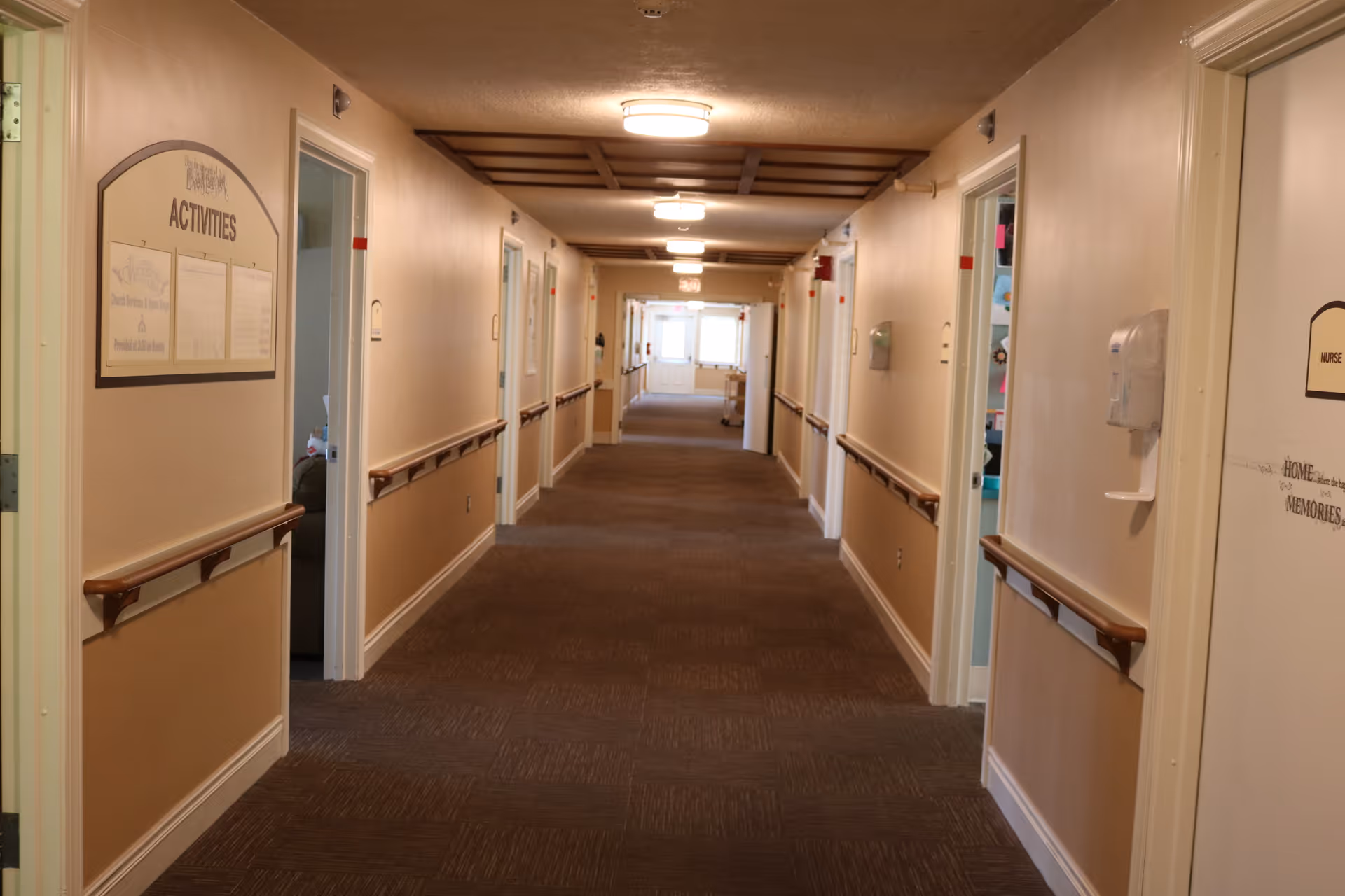A long, well-lit hallway in a senior living facility with beige walls, brown carpet, handrails on both sides, and several open doors leading to rooms. There is a sign labeled 'ACTIVITIES' on the left wall and a door labeled 'NURSE' on the right. The hallway extends to a bright exit or window at the far end.