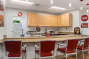 Interior view of a kitchen area with a granite countertop bar and five red cushioned bar stools. Light wood cabinets are mounted on the wall above the countertop. The kitchen includes a refrigerator, microwave, and a coffee maker. The walls are decorated with vintage-style Coca-Cola signs and a small plant is placed on top of the refrigerator. The ceiling has recessed lighting and pendant lights above the bar.