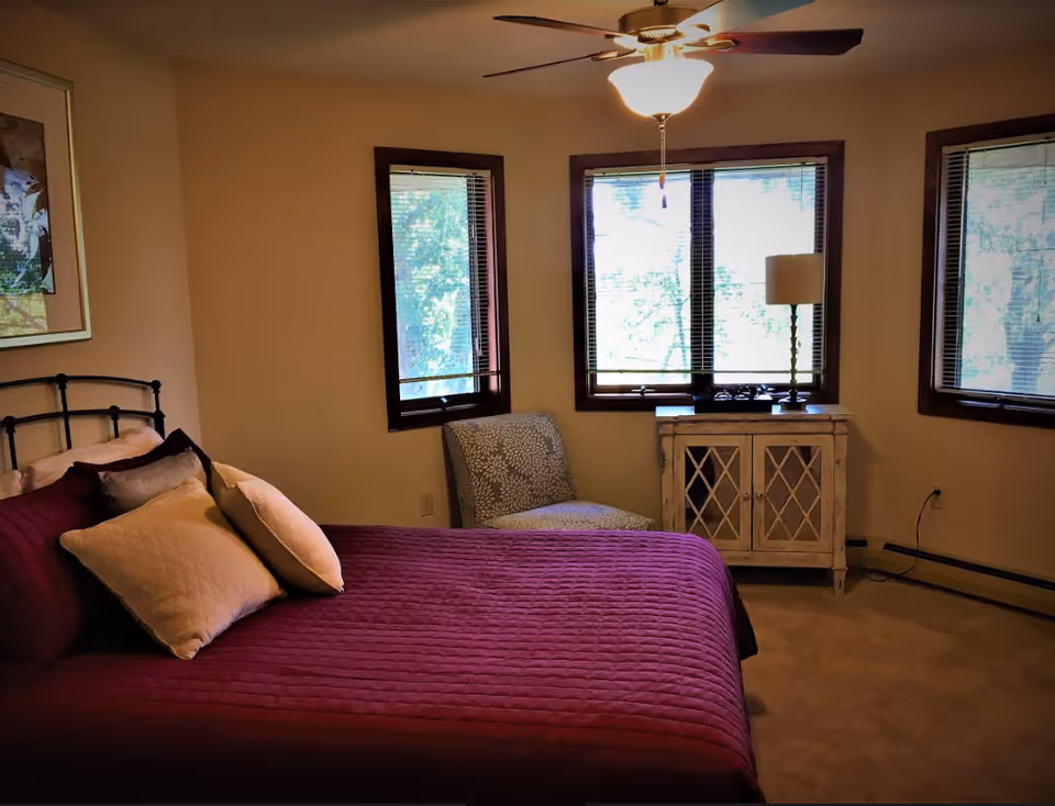 A bedroom with a bed covered in a purple quilt and several pillows, a patterned chair near three windows with blinds, a small white cabinet with a lamp on top, and a ceiling fan with a light.