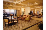 A spacious and elegantly furnished living room area with multiple seating arrangements including armchairs and sofas. The room features large windows with a view of the outdoors, patterned carpet flooring, and warm lighting from ceiling fixtures. There are decorative plants and flowers placed throughout the room.
