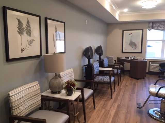 Seating area in a senior living facility with chairs, side tables, wall art and salon hair-dryers.