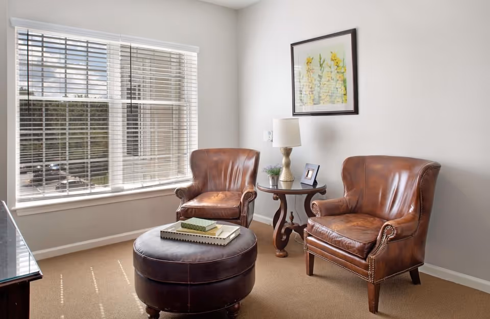 Two brown leather armchairs and a round ottoman arranged by a window in a bright sitting area with a side table and lamp.