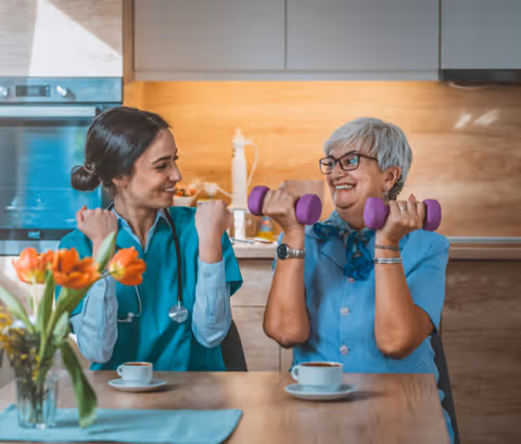 A young female healthcare worker and an elderly woman with gray hair and glasses are sitting at a kitchen table, smiling and lifting purple dumbbells together. There are two cups of coffee on the table and a vase with orange tulips in the foreground.