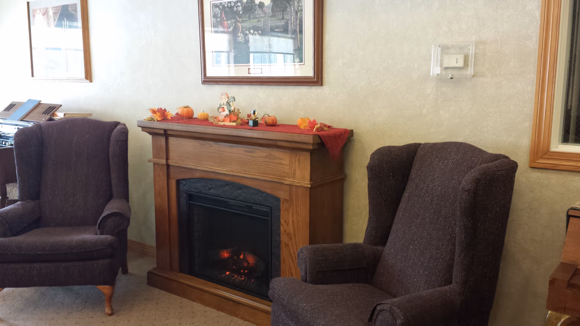 A cozy interior room featuring two dark upholstered wingback chairs positioned on either side of a wooden electric fireplace. The fireplace mantel is decorated with small autumn-themed items including pumpkins and leaves. Above the fireplace hangs a framed picture. The room has light-colored walls and carpeted flooring.