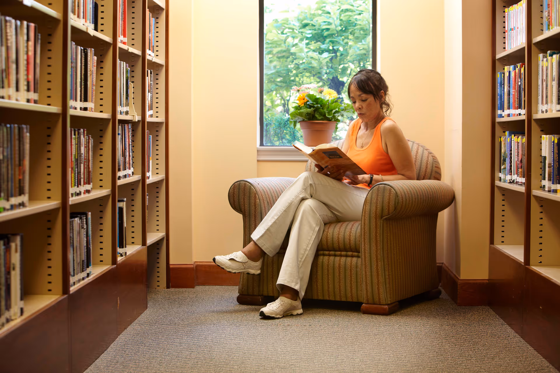 A woman in an orange sleeveless top and white pants is sitting on a striped armchair reading a book in a library. She is positioned between two tall bookshelves filled with books, and there is a window behind her with a potted plant on the windowsill.