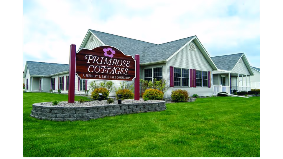 Exterior view of Primrose Cottages, a memory and basic care community, showing a single-story building with light-colored siding, purple shutters, and a well-maintained green lawn with a stone-bordered flower bed and a wooden sign in front.