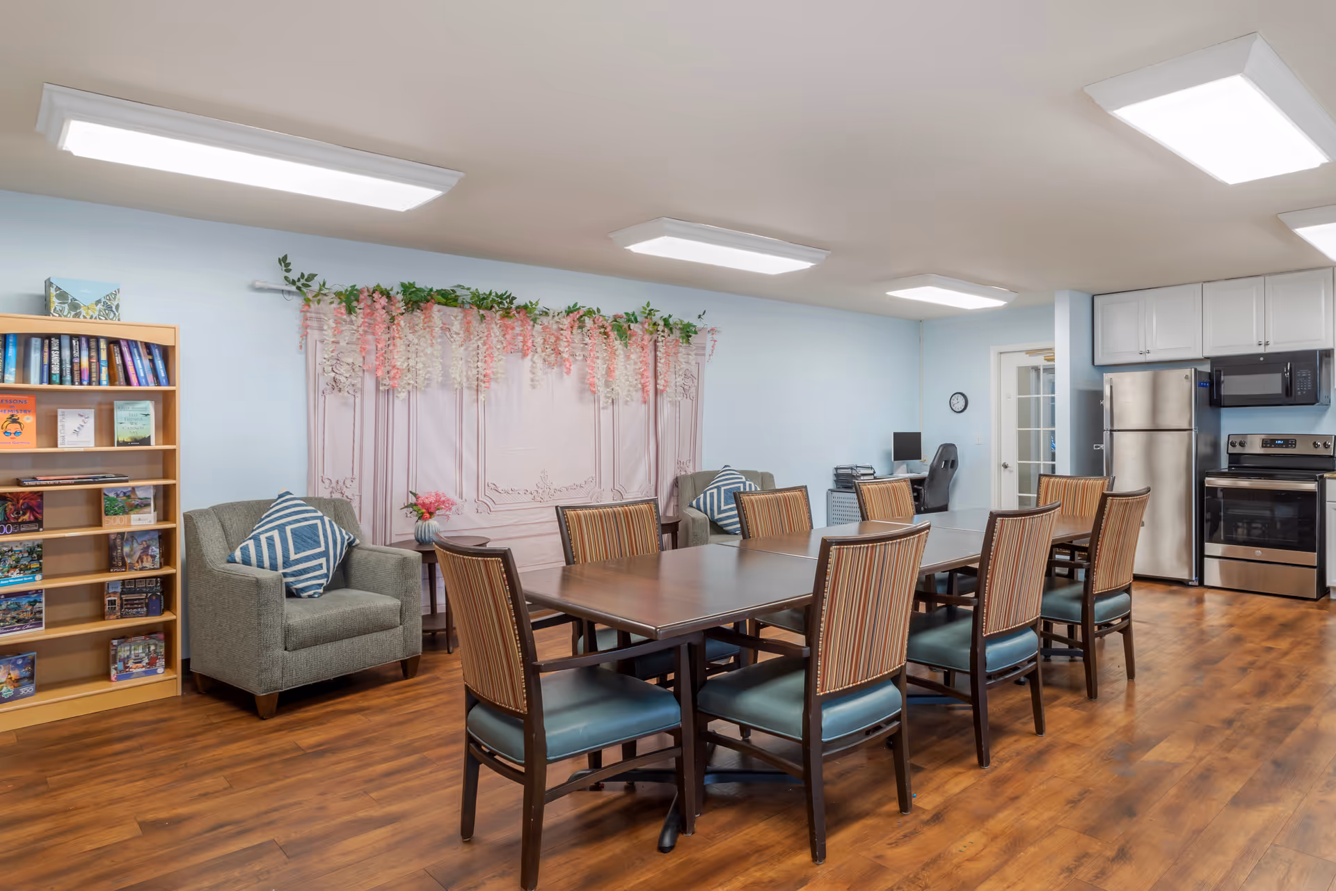 A communal dining and activity room with a long wooden table surrounded by eight chairs with striped upholstery. The room has light blue walls, wood flooring, and bright ceiling lights. There is a small kitchen area with a stainless steel refrigerator, stove, and microwave. Two gray armchairs with blue and white patterned pillows are placed near a bookshelf filled with books and puzzles. A decorative pink and white floral arrangement hangs on the wall behind the armchairs.
