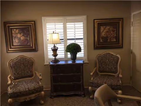 Two upholstered chairs flank a wooden cabinet with a lamp, plant, and framed art.