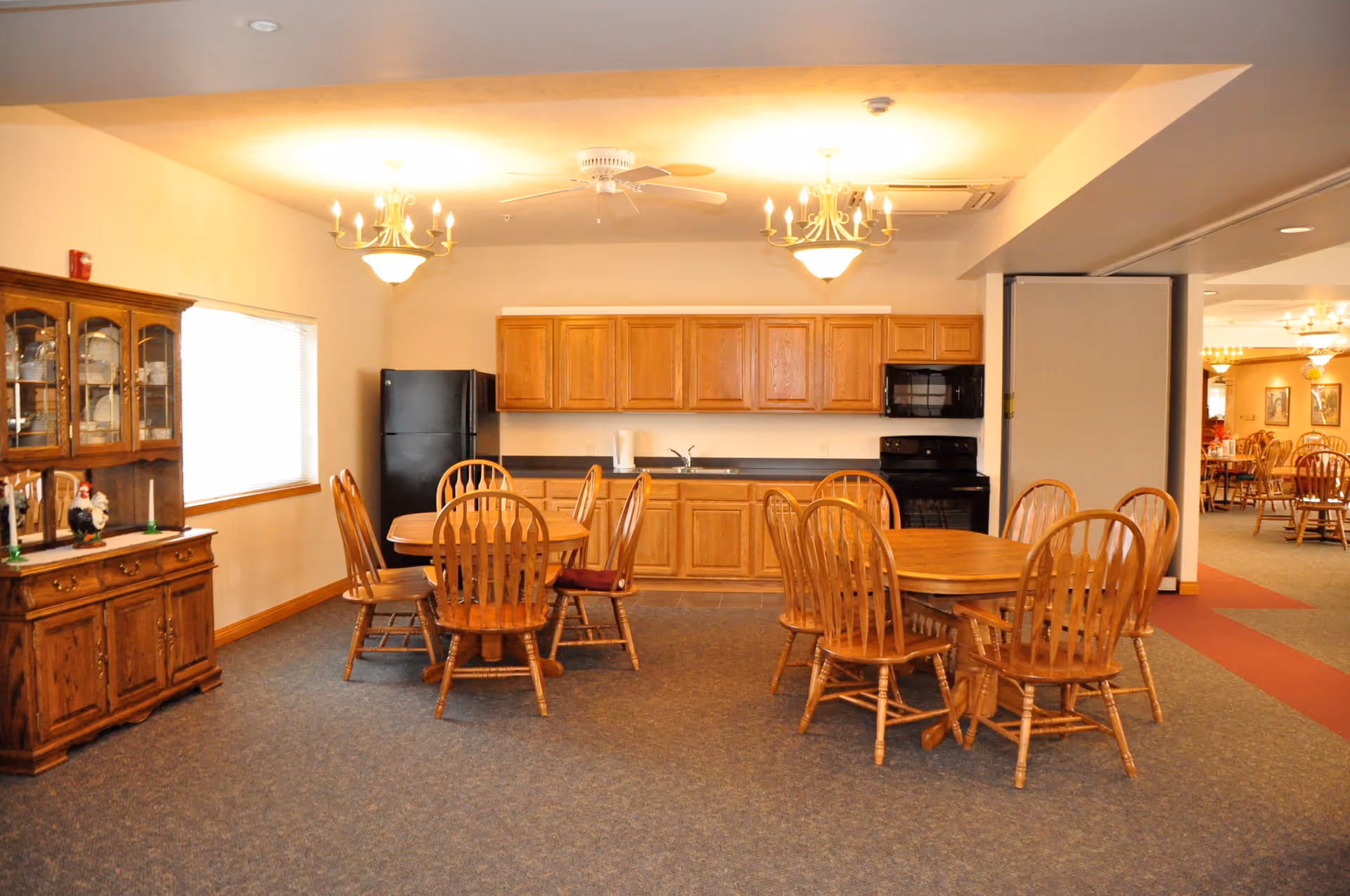 Communal dining area with wooden tables and chairs in front of a small kitchen with oak cabinets and appliances.