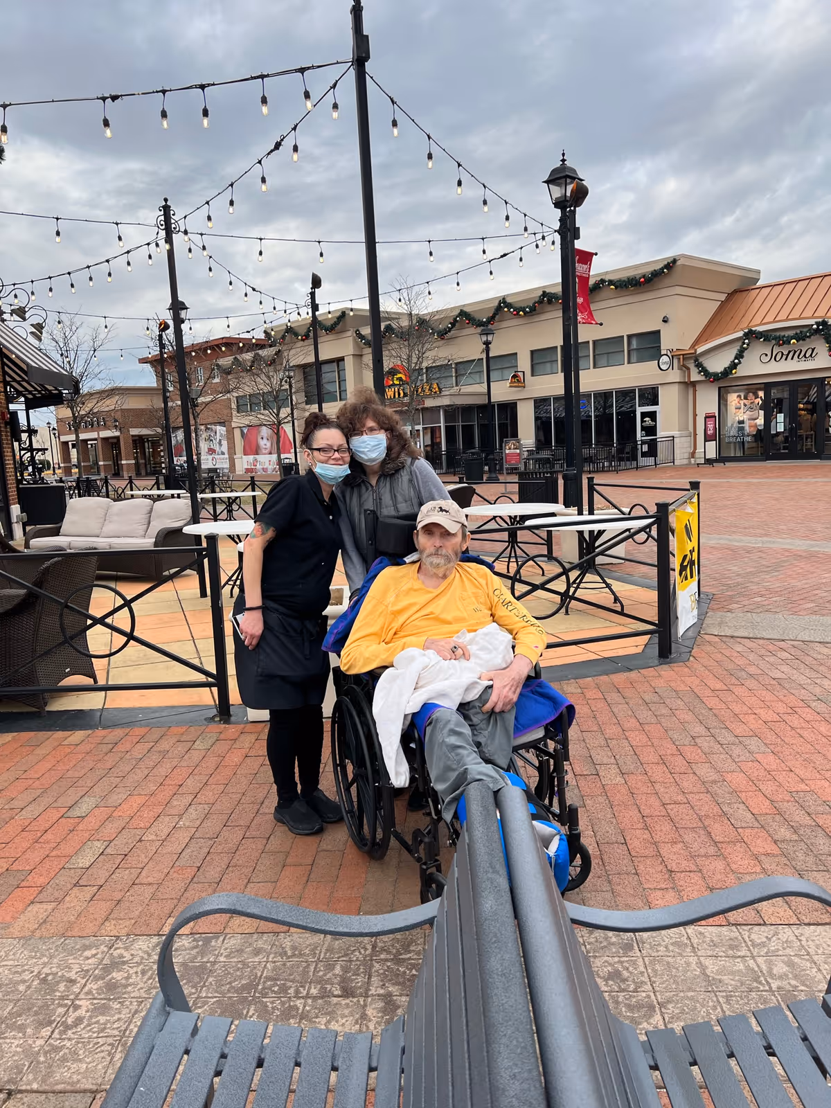 Three people outdoors in a shopping plaza with brick pavement and string lights overhead. Two women wearing face masks stand behind a man in a wheelchair who is wearing a yellow shirt and a beige cap. There are outdoor seating areas and storefronts decorated with garlands in the background under a cloudy sky.
