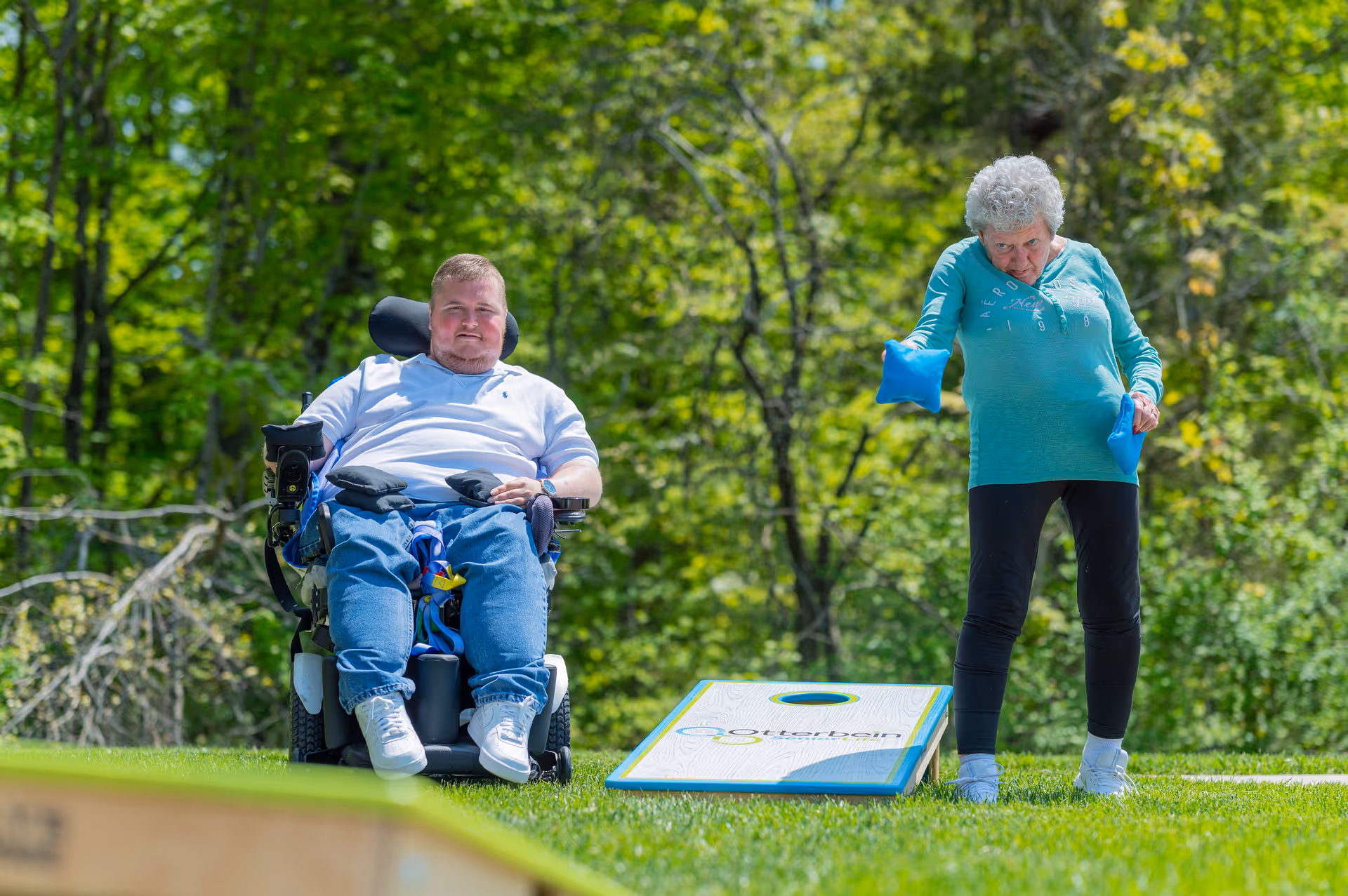 An elderly woman playing cornhole outdoors, tossing bean bags towards a cornhole board on the grass, while a man in a motorized wheelchair watches nearby. The background shows green trees and a sunny day.