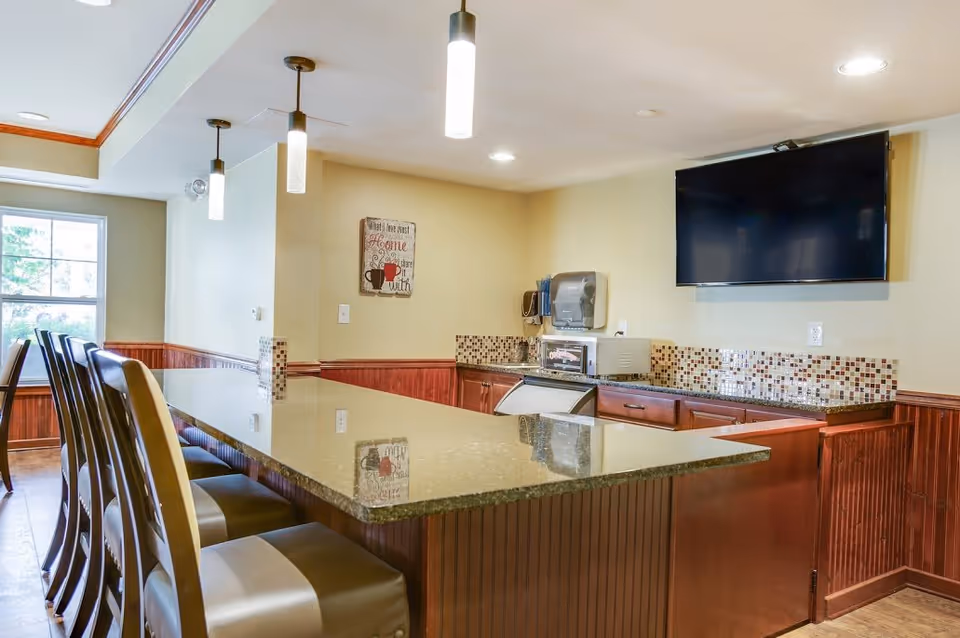 Interior view of a kitchen area with a long granite countertop bar and several high chairs. The kitchen has wooden cabinets, a mosaic tile backsplash, a mounted flat-screen TV, and pendant lights hanging from the ceiling. A window lets in natural light from the left side.