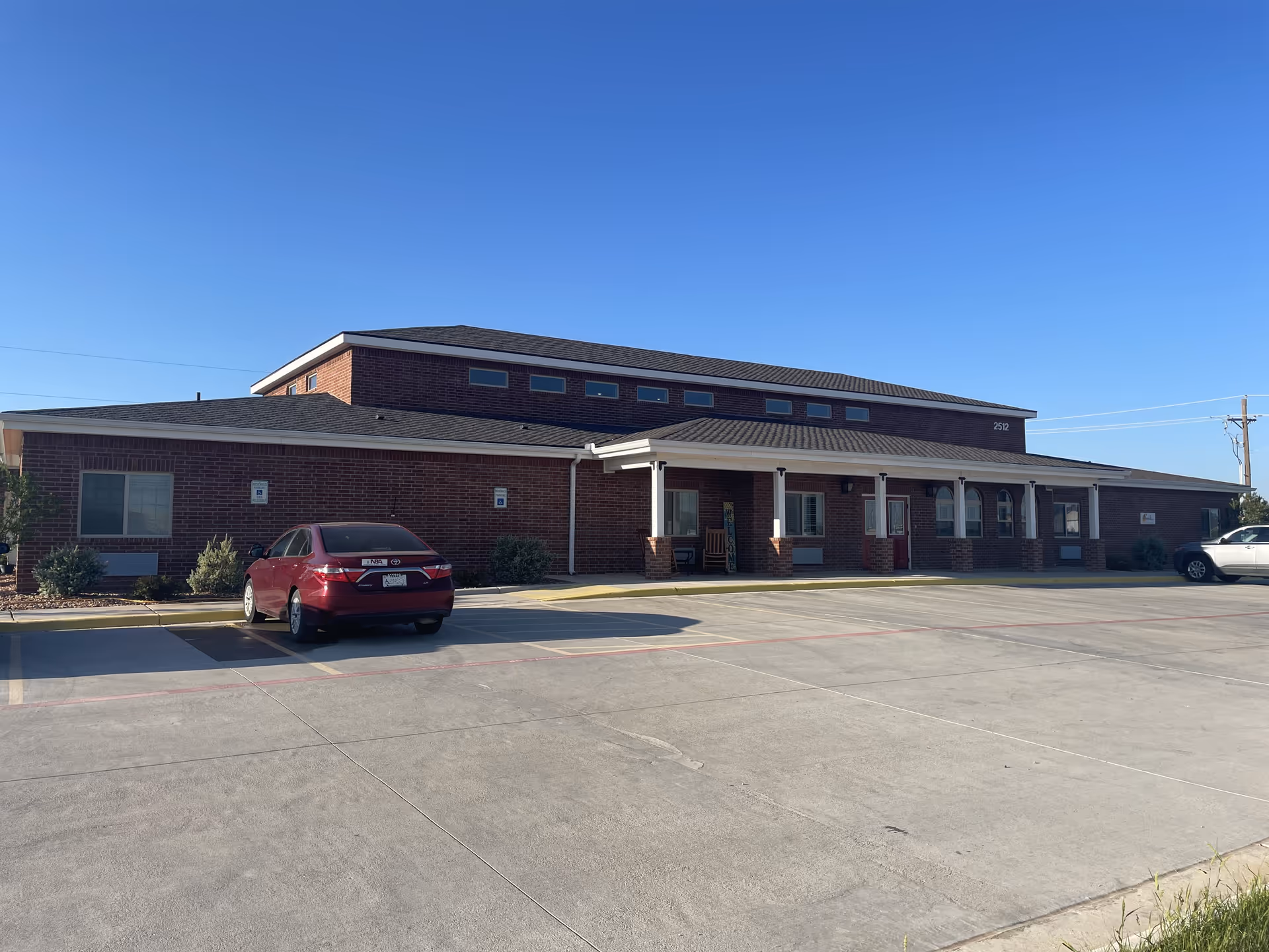 Exterior view of a single-story brick building with a covered entrance and a parking lot in front. Two cars are parked in the lot under a clear blue sky.