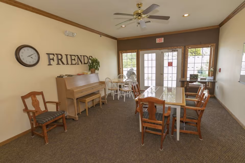 A cozy common room with a piano, several wooden chairs, and tables. The wall has a clock and the word 'FRIENDS' displayed. There are large windows and a glass door letting in natural light, and a ceiling fan is mounted above.