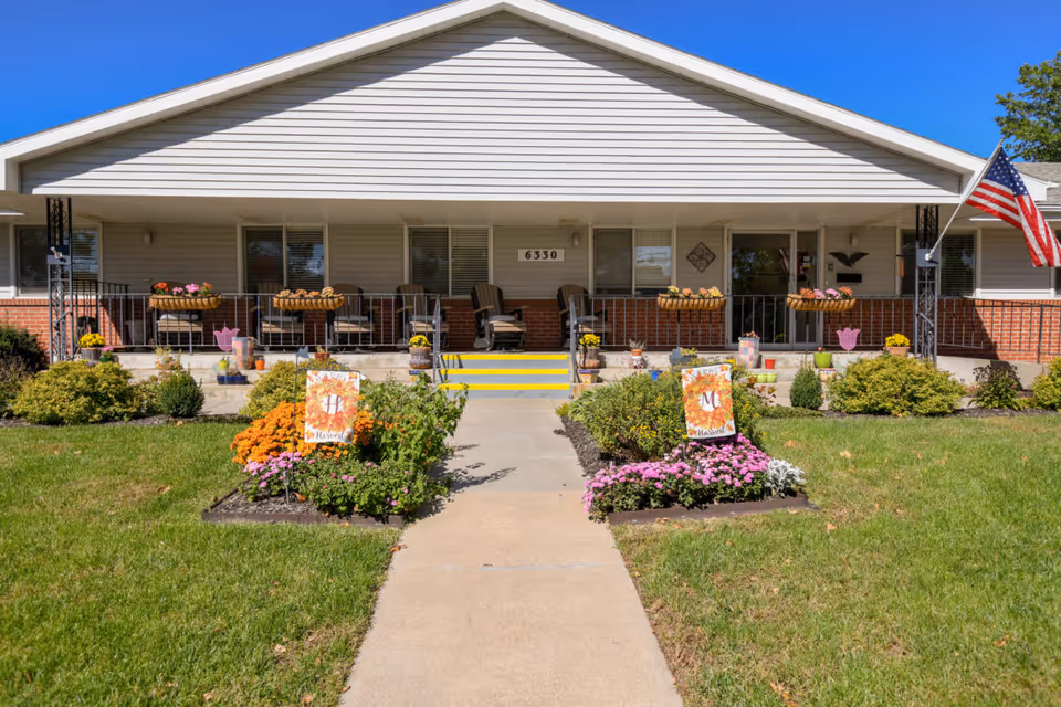 Front exterior view of a single-story building with a covered porch, several chairs, flower pots, and two flower beds with decorative signs. An American flag is displayed on the right side of the porch. The building number 6330 is visible above the steps.