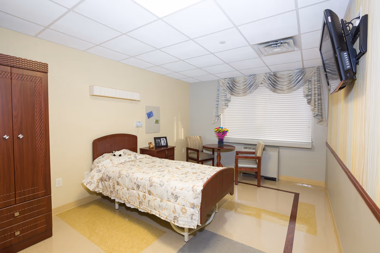 A single bed with floral bedding in a senior living facility room. The room has a wooden wardrobe, a small wooden nightstand with framed photos, a round table with two chairs, a vase of colorful flowers, a window with blinds and decorative curtains, and a wall-mounted TV.