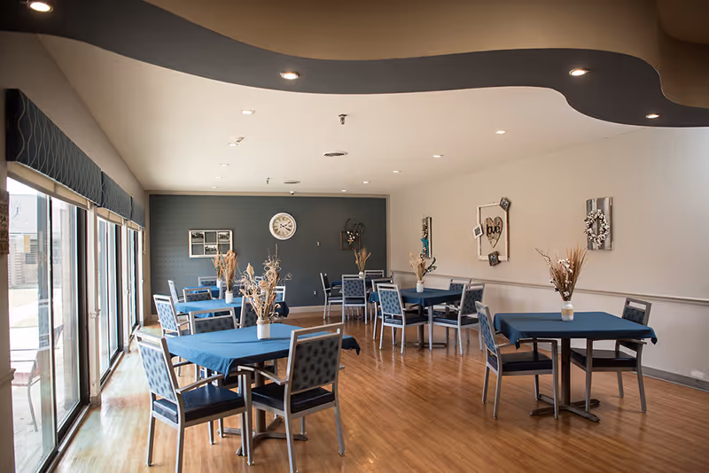 A dining room with several tables covered with blue tablecloths and decorated with vases of dried flowers. The room has wooden flooring, large windows on one side with blue valances, and a dark accent wall with a clock and framed decorations. The ceiling has recessed lighting and a curved design element.