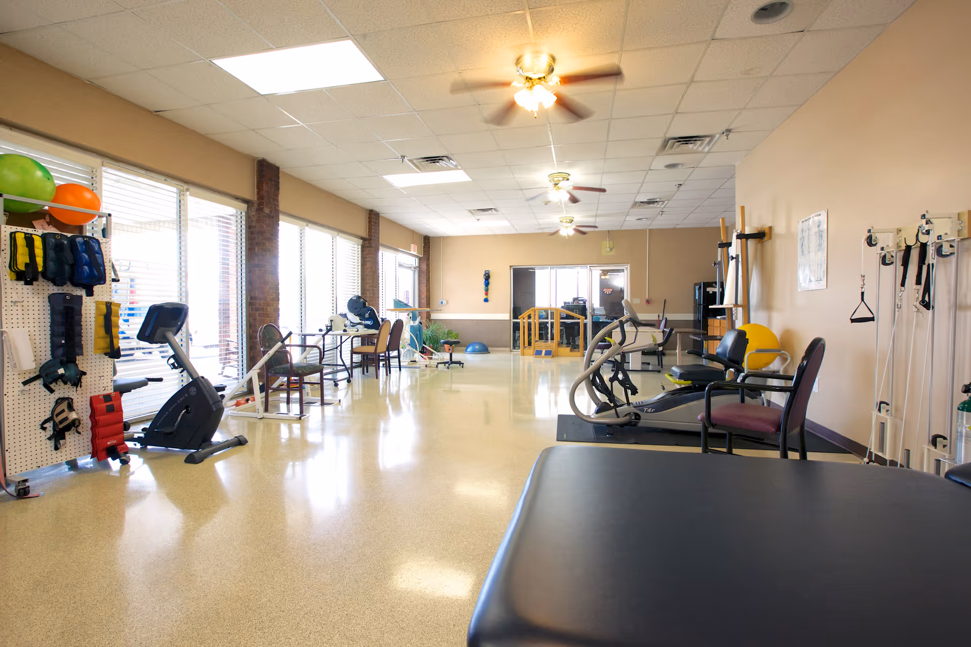 A spacious rehabilitation or physical therapy room with exercise equipment including stationary bikes, therapy balls, and various rehabilitation tools. The room has large windows on one side allowing natural light, ceiling fans, and chairs positioned around the equipment.