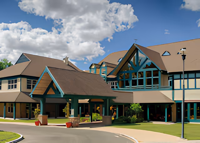 Exterior view of Geer Village Lodge, a large building with multiple peaked roofs and large windows under a partly cloudy sky. The entrance features a covered driveway with wooden beams and stone pillars, surrounded by greenery and a paved road.