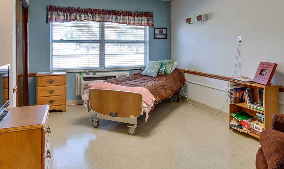 A simple nursing center bedroom with a single hospital-style bed covered with a brown blanket and two decorative pillows. The room has a large window with blinds and a plaid valance, a wooden dresser, a small bookshelf filled with books and games, and a brown armchair partially visible. The walls are painted light blue and white, and the floor is covered with beige linoleum tiles.