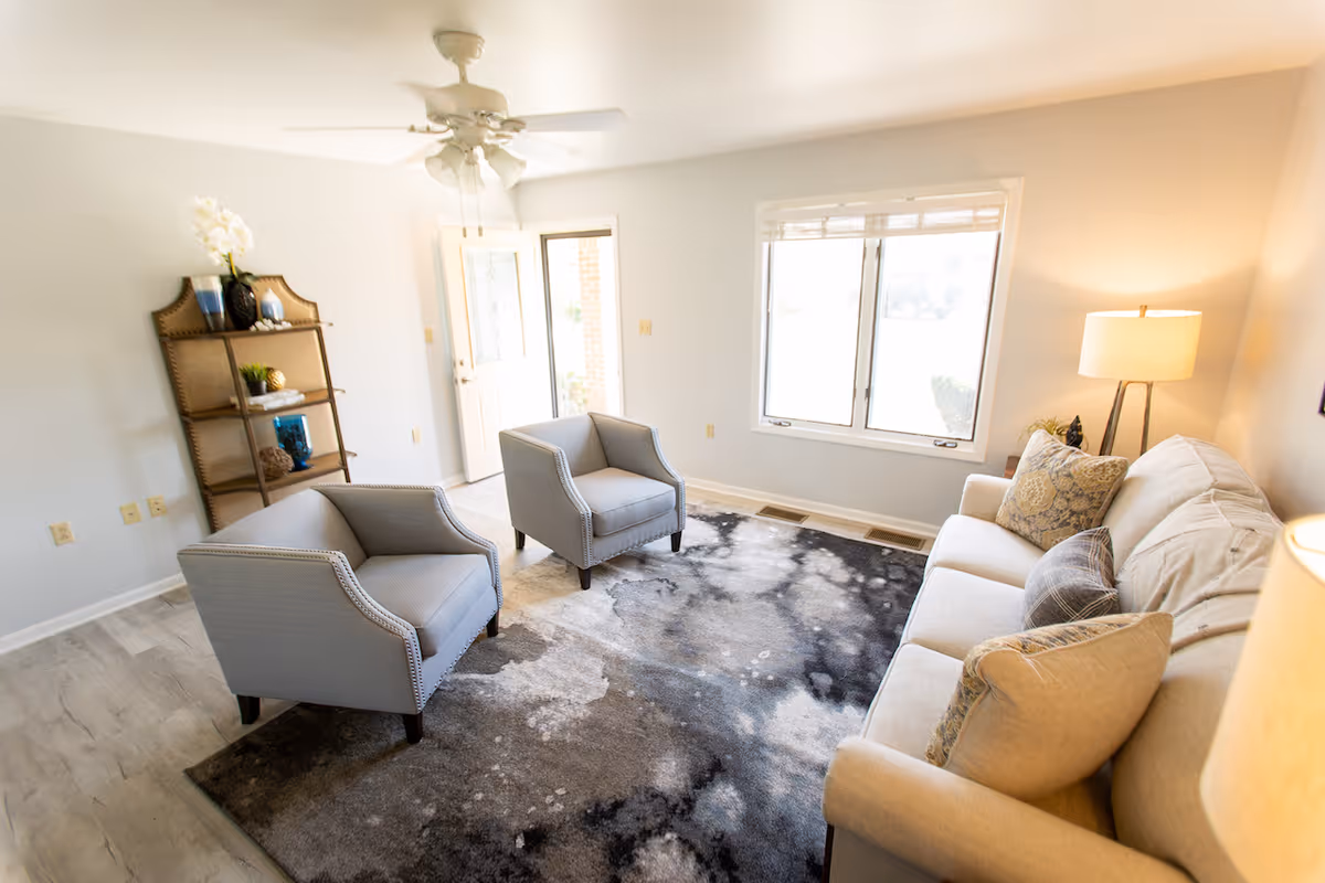 A bright living room with a beige sofa adorned with multiple pillows, two gray armchairs, a patterned area rug, a wooden shelving unit with decorative items, a ceiling fan, and a window letting in natural light.