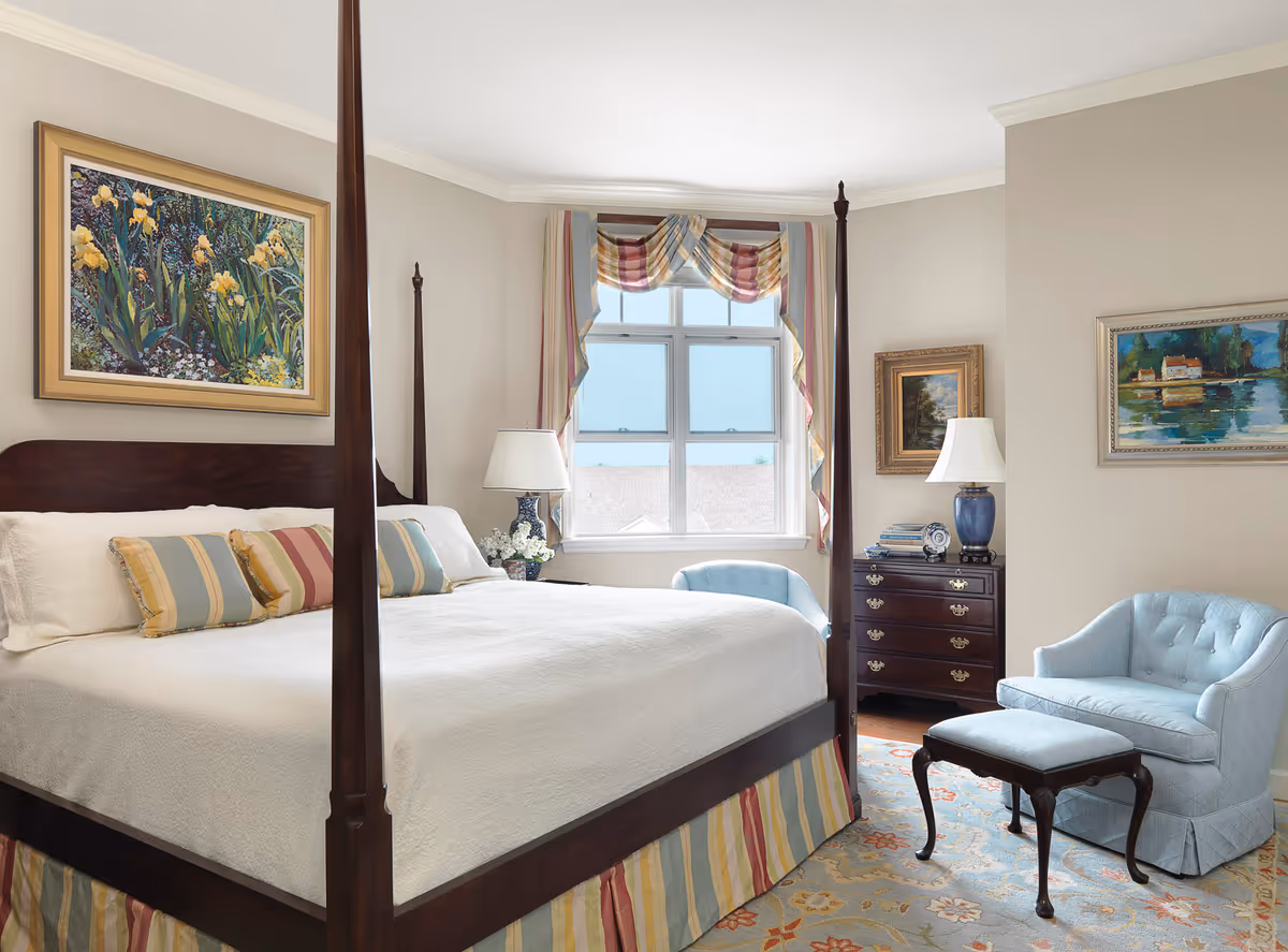 A bright and elegant bedroom featuring a large four-poster bed with white bedding and striped pillows. The room has a window with colorful drapes, two blue upholstered chairs with a matching ottoman, a dark wooden dresser with a lamp and books, and framed paintings on the walls.