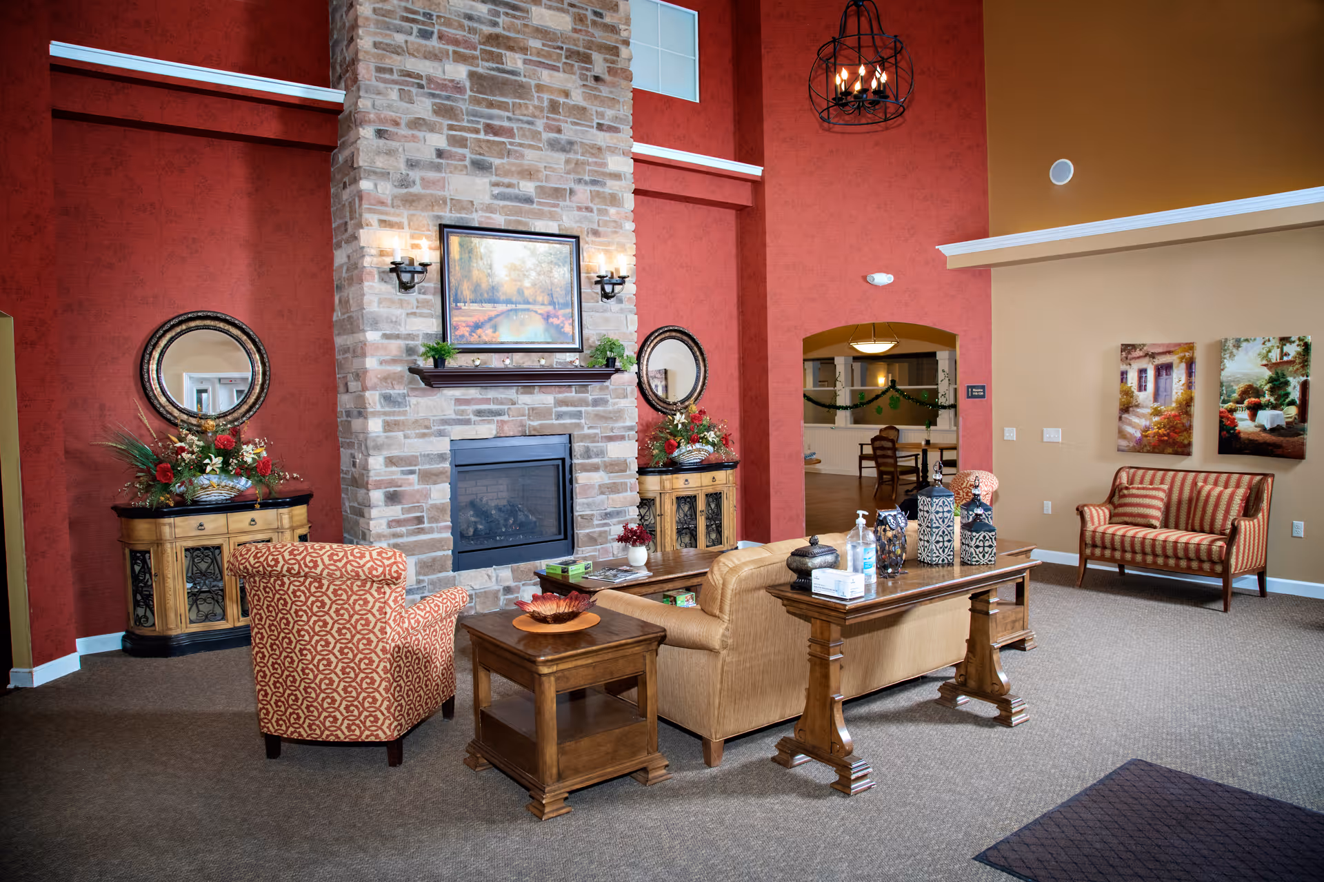A cozy living room area with a stone fireplace in the center, flanked by two wooden cabinets with round mirrors and floral arrangements. There is a patterned armchair, a beige sofa, and a wooden coffee table with decorative items. The walls are painted in warm red and beige tones, and there are two landscape paintings on the right wall.