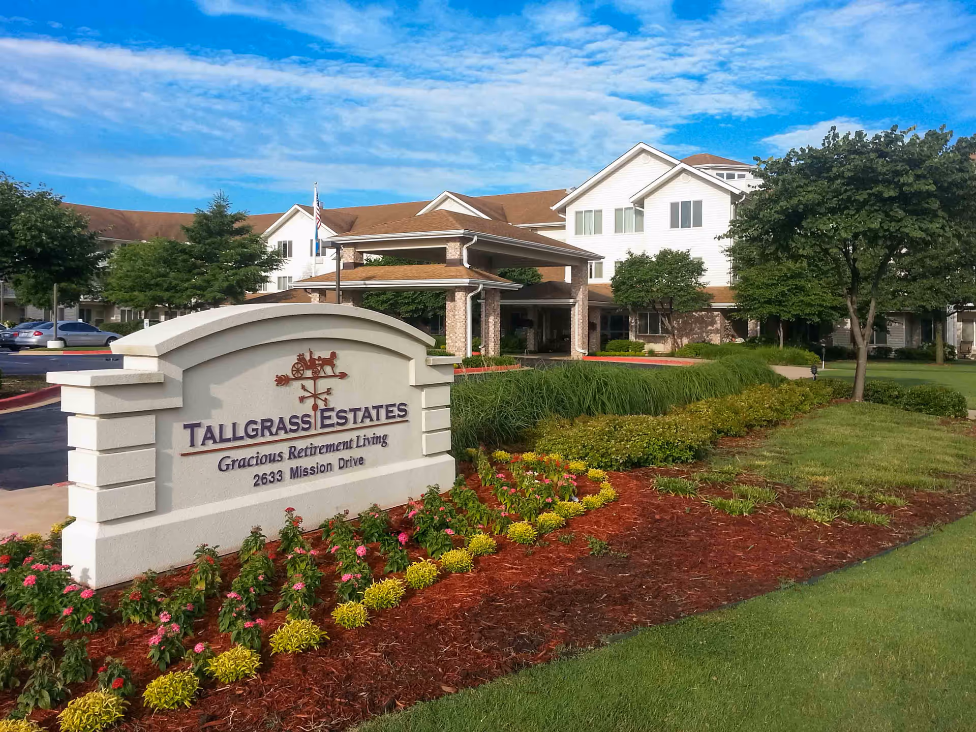 Exterior view of Tallgrass Estates retirement living facility with a large sign in the foreground displaying the name and address. The building has a covered entrance, multiple windows, and is surrounded by well-maintained landscaping including trees, bushes, and flower beds under a partly cloudy blue sky.