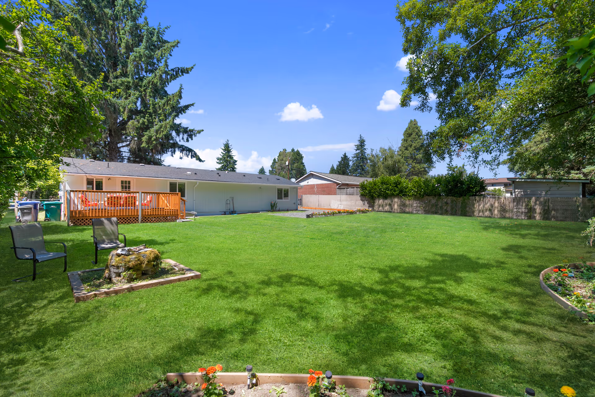 A spacious, well-maintained backyard with lush green grass, a small garden bed with flowers in the foreground, two black chairs around a small stone fire pit, and a wooden deck attached to a single-story white building. Tall trees and a clear blue sky with a few clouds are visible in the background.
