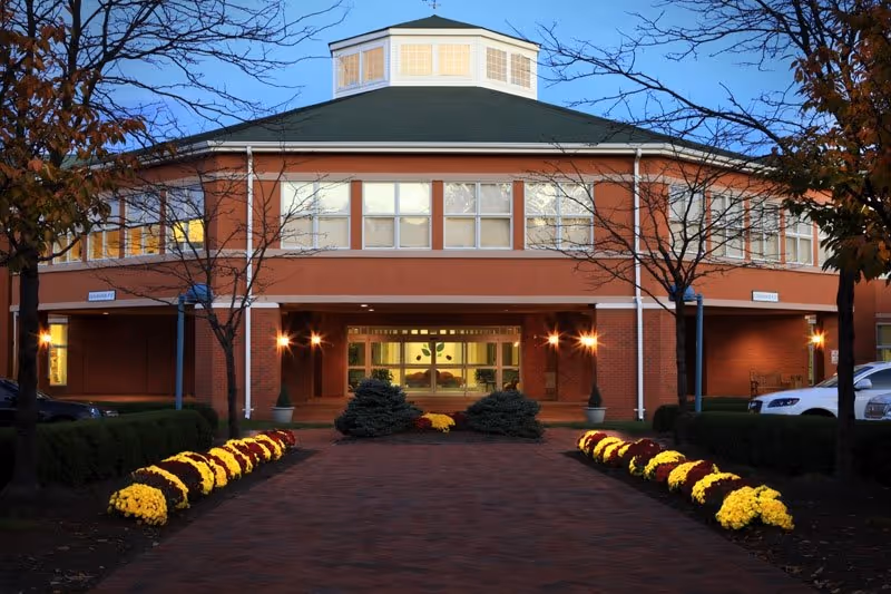 Front entrance of a circular brick building with a green-roofed cupola, illuminated walkway, and rows of yellow flowers.