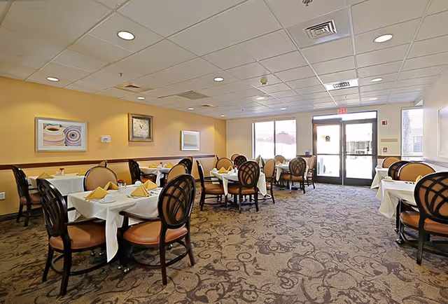 A dining room in a senior living facility with multiple tables covered in white tablecloths, each set with folded yellow napkins and small flower vases. The room has patterned carpet flooring, yellow walls with framed artwork, and large windows letting in natural light near the exit door.