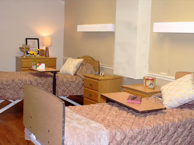 A room with two hospital-style beds covered with patterned bedspreads and pillows. Each bed has an overbed table, one with a remote control and a book on it. There are two wooden nightstands between and beside the beds, with decorative items including a small clock, a flower vase, and a framed quote. The walls are painted in neutral tones, and the floor is wooden.
