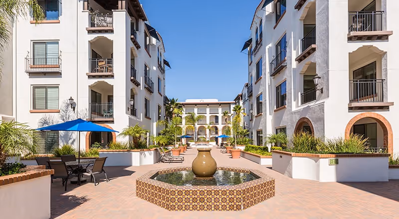 Outdoor courtyard area of a senior living facility with white multi-story buildings on both sides, a central decorative water fountain with patterned tiles, potted plants, patio tables with blue umbrellas, and clear blue sky overhead.