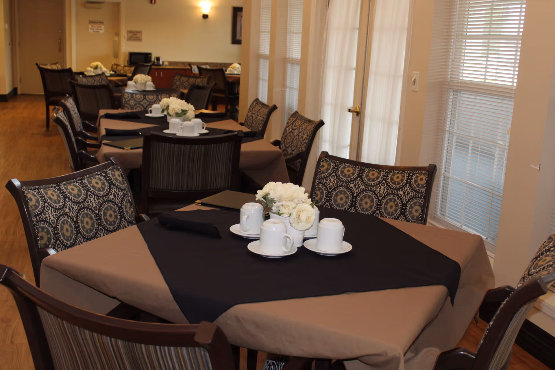 Dining area with multiple tables set with cups and floral centerpieces and patterned chairs by windows.