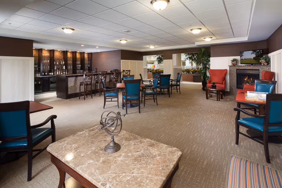 Spacious senior living common area with tables and chairs, a bar, and a fireplace.