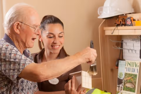 An elderly man and a younger woman are engaged in a craft activity. The man is painting a small wooden box with a brush while the woman looks on and smiles. They are indoors, with shelves holding various items including a hard hat and books in the background.