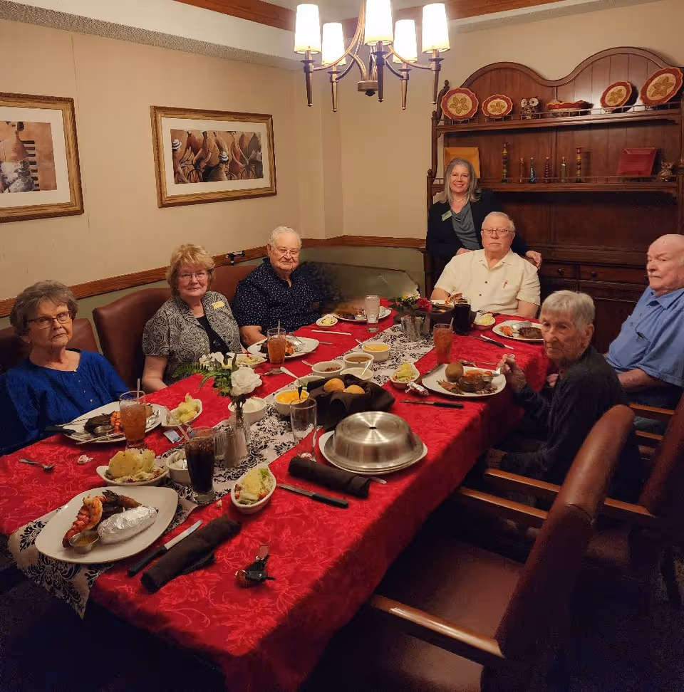 A group of elderly people seated around a dining table with a red tablecloth, enjoying a meal together in a warmly lit room. A woman stands behind the group, smiling. The table is set with plates of food, drinks, and decorative flowers. The room has framed artwork on the walls and a wooden cabinet with decorative plates and items.
