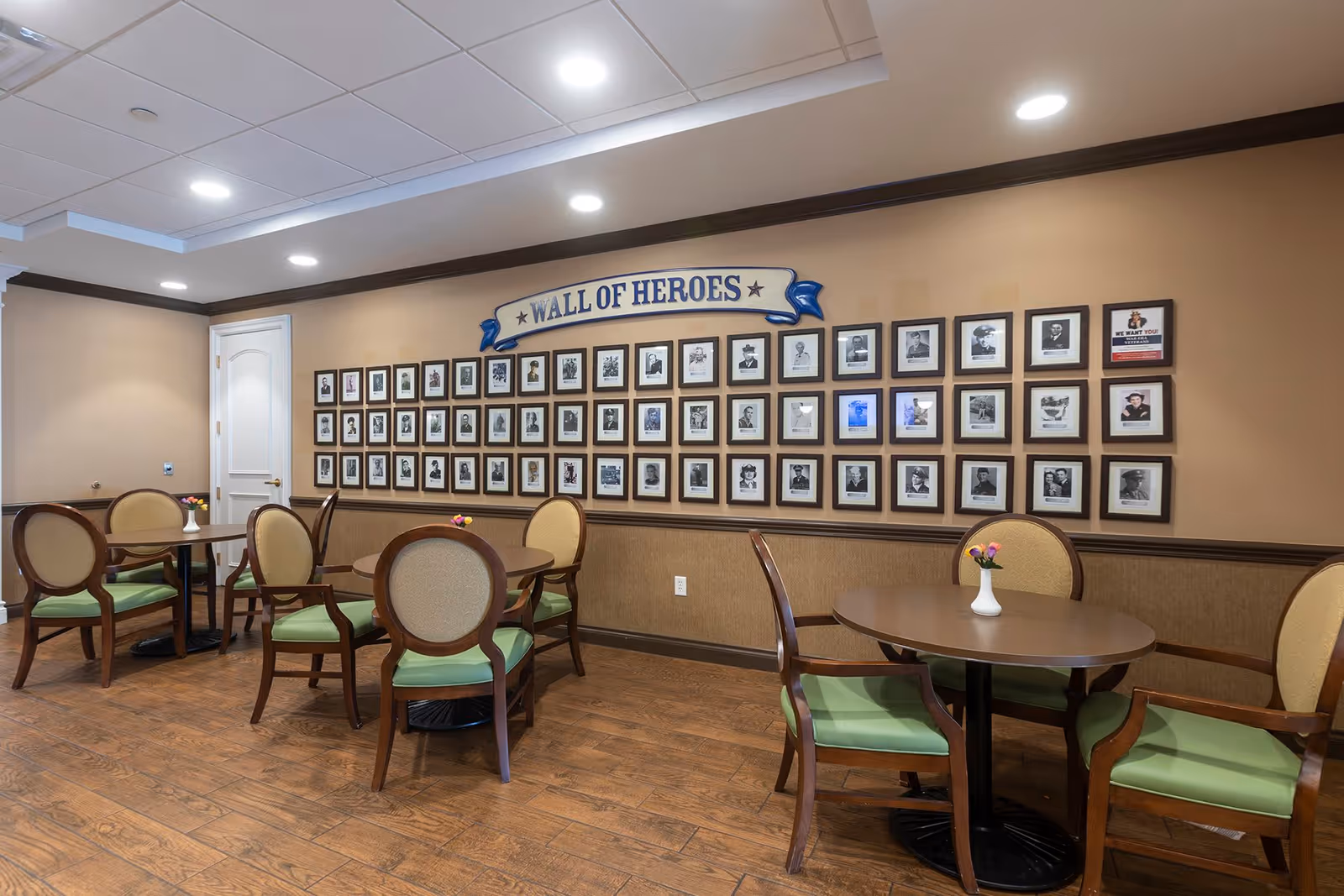 A room with several round tables and chairs with green cushions, a wooden floor, and a wall displaying a 'Wall of Heroes' featuring framed photographs of various individuals.