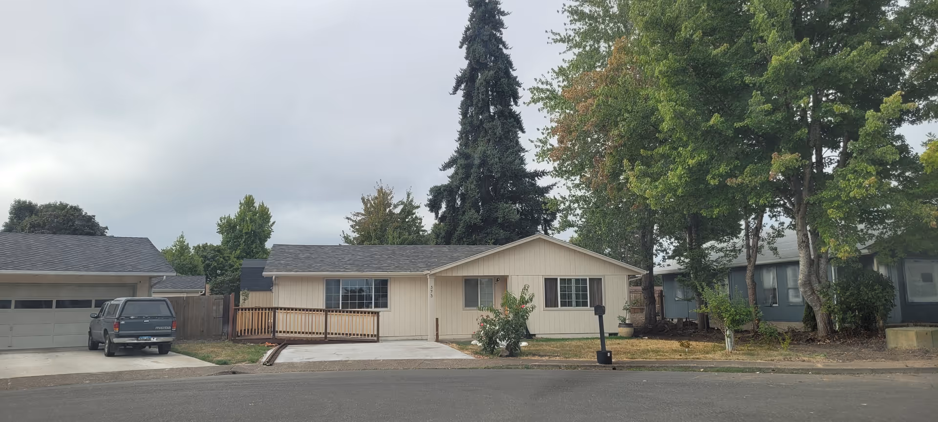 A single-story beige residential building with a small front porch and driveway, flanked by neighboring homes and tall trees.