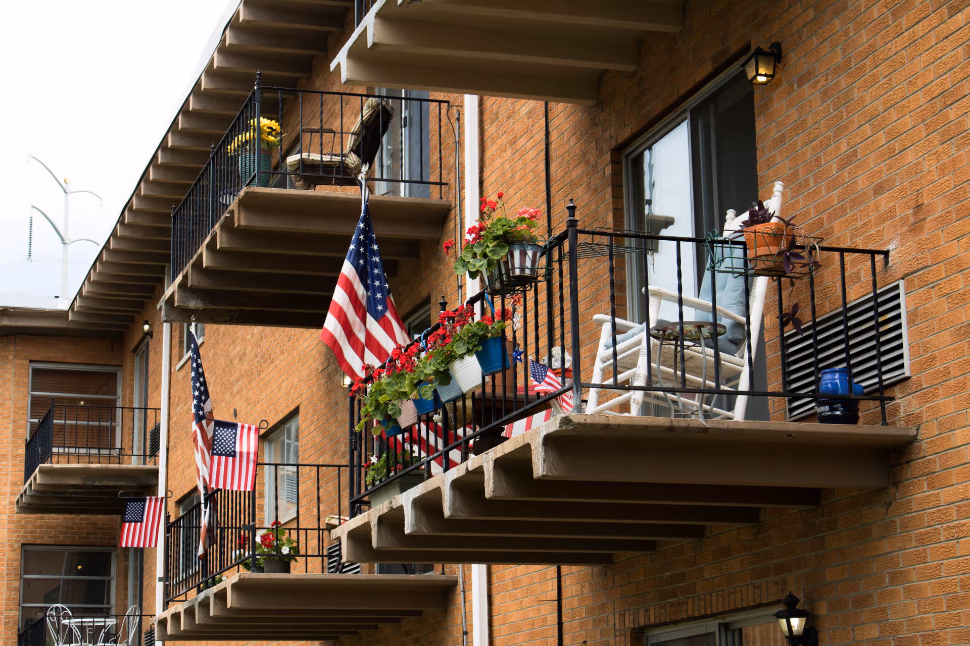 Exterior view of a brick building with multiple balconies decorated with American flags, potted plants, and white rocking chairs.