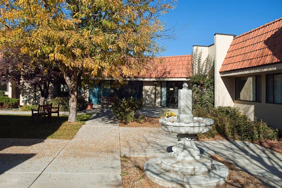 Outdoor courtyard area of a senior living facility with a stone fountain in the foreground, surrounded by bushes and trees with autumn foliage. The building has beige walls and red-tiled roofs with large windows. There are benches placed along the walkway.