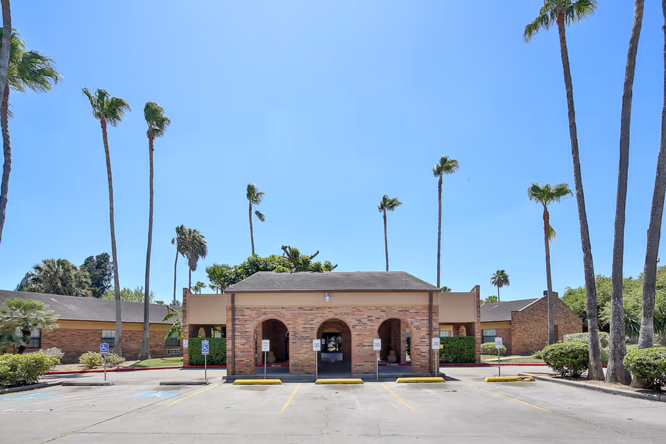 Exterior view of Ebony Lake Nursing and Rehabilitation Center showing a brick building with three arched entrances, surrounded by tall palm trees and clear blue sky. There is a parking lot with several handicap parking spaces in front of the building.