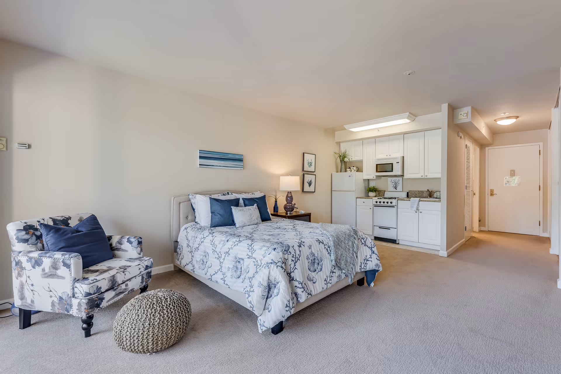A studio apartment interior featuring a bed with blue and white patterned bedding, a floral armchair with a navy pillow, a knitted pouf, and a small kitchen area with white cabinets, a refrigerator, stove, and microwave. The room has beige walls and carpet, with a hallway leading to a white door.