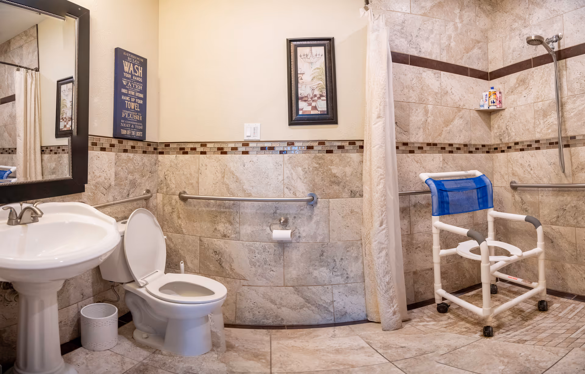 A bathroom with beige tiled walls and floor, featuring a white pedestal sink, a toilet with the lid open, a wall-mounted mirror, and a shower area with a white curtain and a white shower chair with blue backrest. There are grab bars installed on the walls for accessibility, a small trash can next to the toilet, a framed picture on the wall, and a decorative sign with bathroom rules.