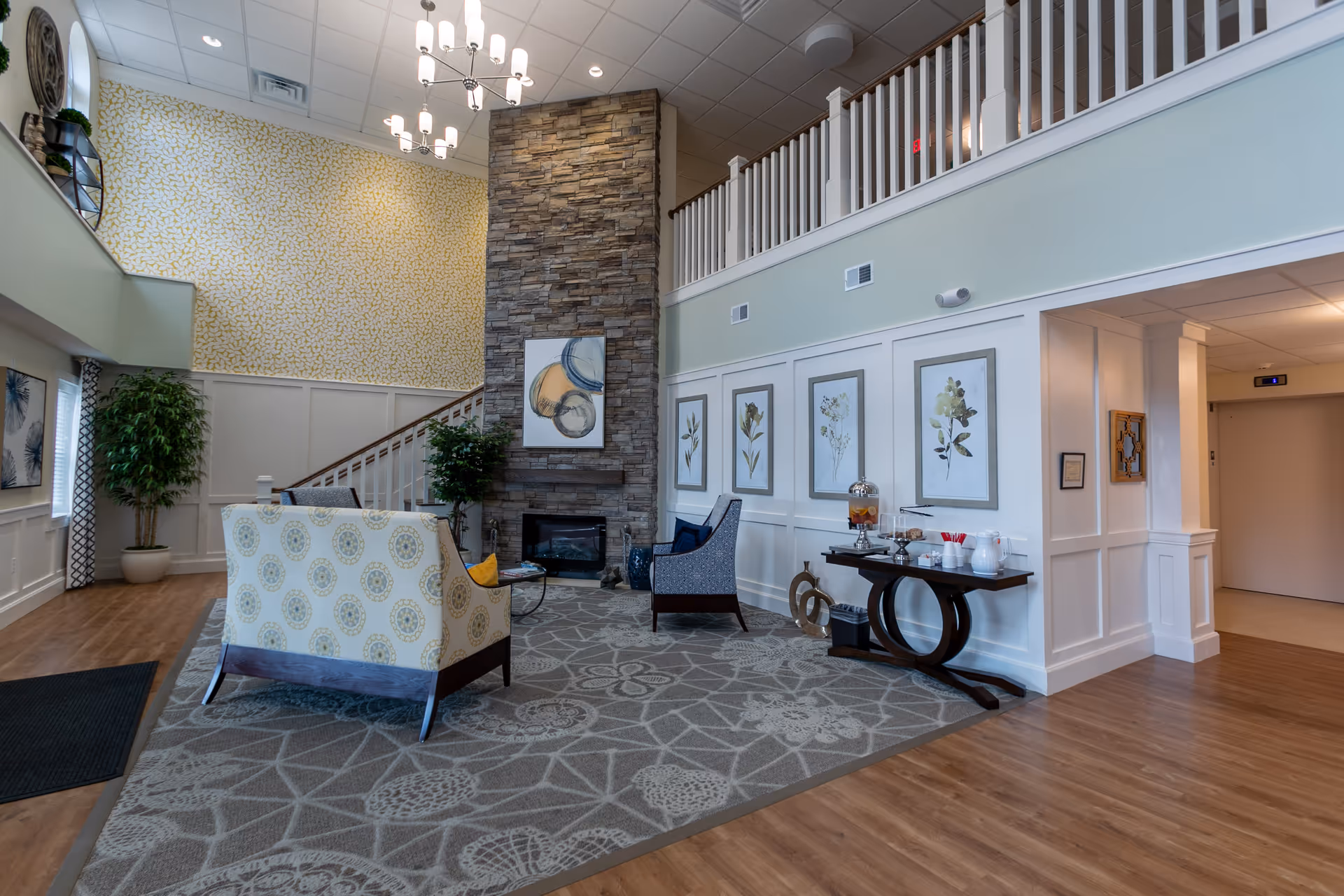 Bright two-story senior living lobby with seating, a stone fireplace, staircase and decorative wall art.