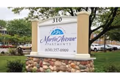 Outdoor image showing a sign for Martin Avenue Apartments with the address number 310 and a contact phone number, surrounded by grass, landscaping rocks, and parked cars in the background.