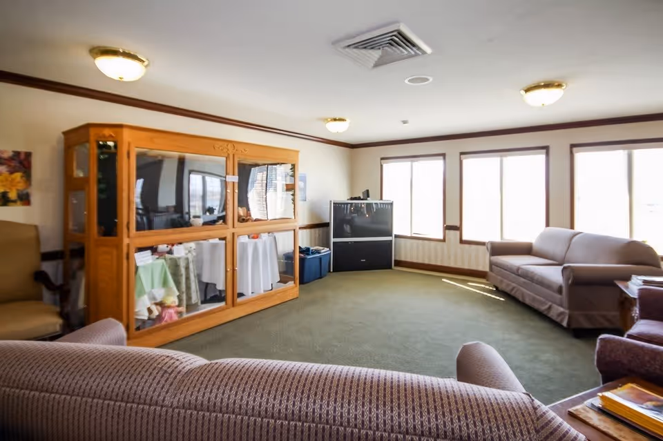 A living room with multiple sofas and armchairs arranged around a large wooden cabinet with glass doors. The room has green carpet, cream-colored walls with wooden trim, and three large windows letting in natural light. A television is placed in the corner near the windows.