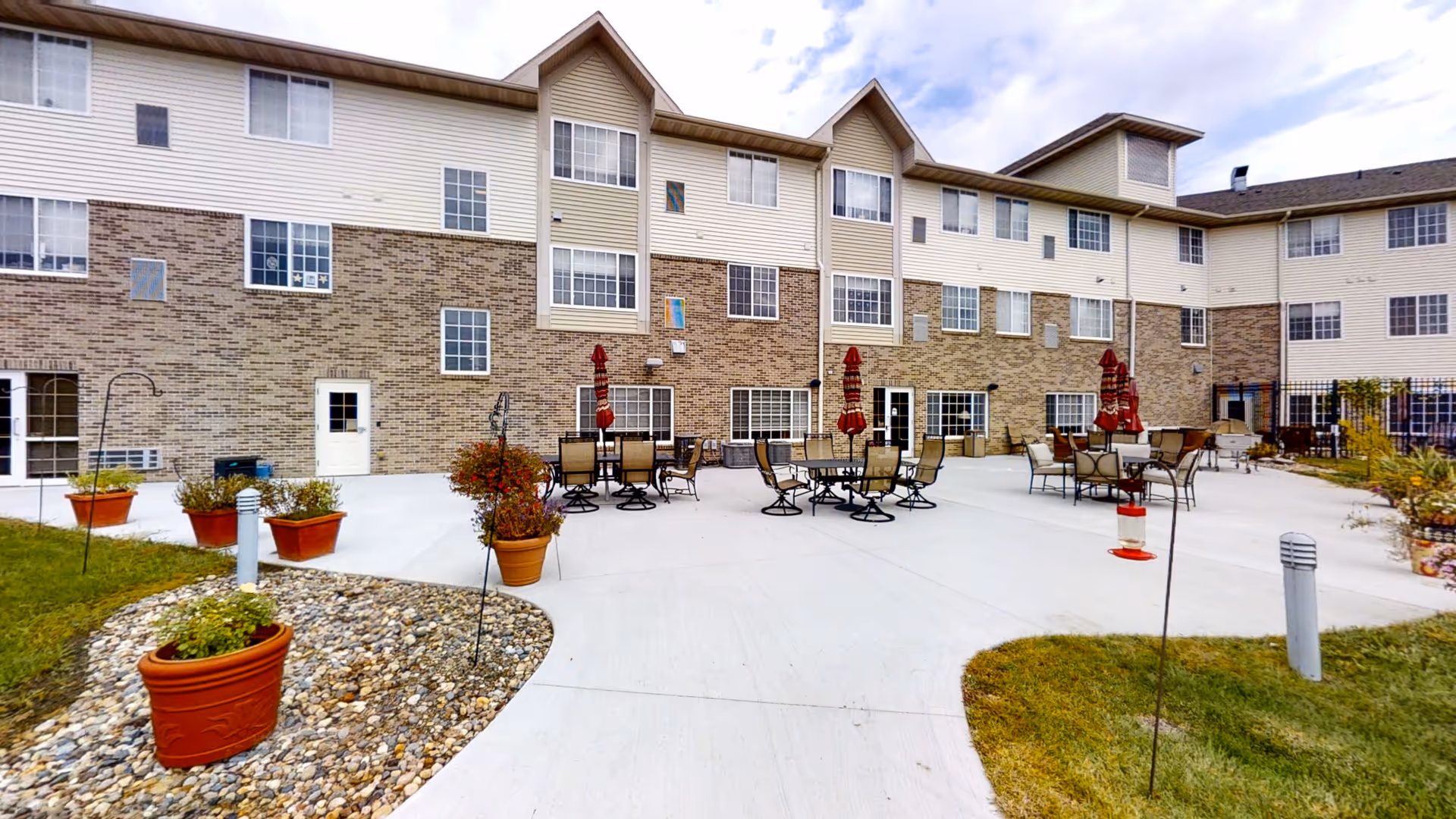 Outdoor patio courtyard with tables, chairs, umbrellas and potted plants in front of a multi-story senior living building.