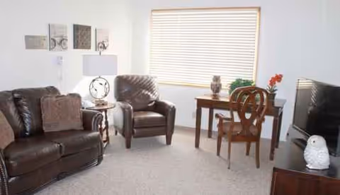 Cozy senior living room with brown leather furniture, window blinds, a desk, and decorative wall art.