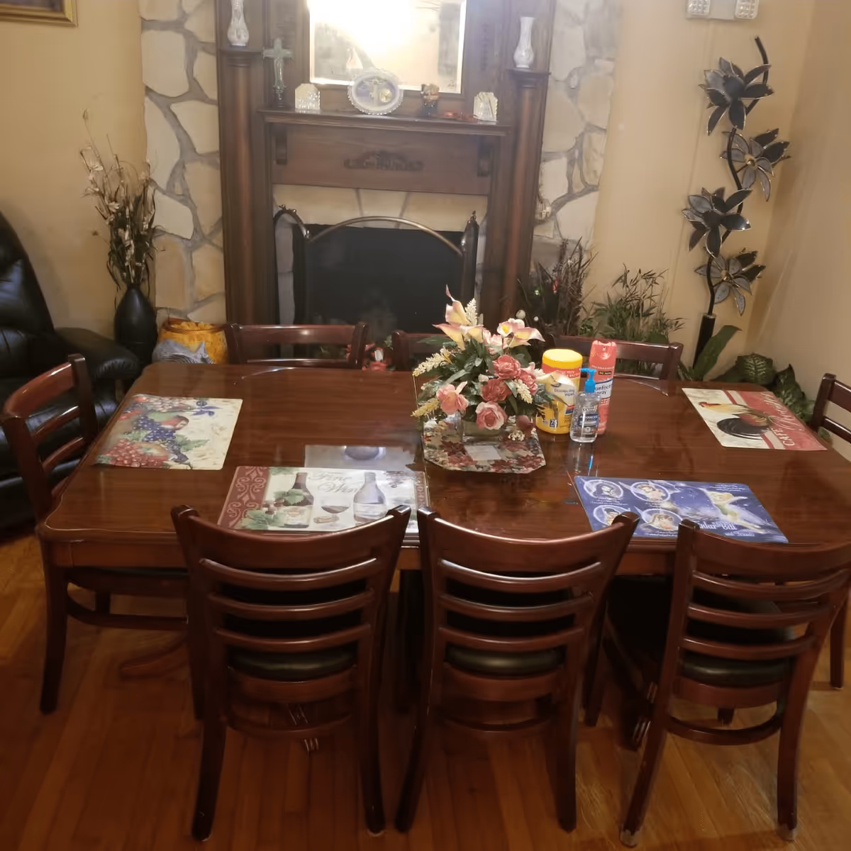 A wooden dining table with six chairs around it, set with various placemats and a floral centerpiece. In the background, there is a stone fireplace with a wooden mantle decorated with small items and a mirror above it. The room also features plants and decorative items on either side of the fireplace.